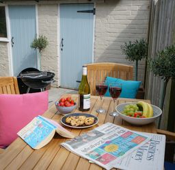 A wooden outdoor table with fruit bowls, cookies, wine glasses, a book, and newspapers on a patio at Smugglers Rest Aldeburgh