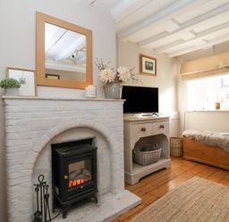 A room with a white brick fireplace and an electric stove inside a wooden floor and a TV on a cabinet near a window with a wooden chest covered by a fur blanket at Rosemary Cottage  Aldeburgh