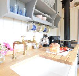 A kitchen countertop with a loaf of bread and tomatoes next to a sink with brass taps and shelves with mugs and bowls above at Shepherds Pightle in Hollesley