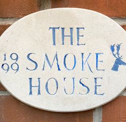 An oval stone plaque with blue carved text reading The Smoke House and the year 1899 on a brick wall at The Smokehouse Cottage in Southwold