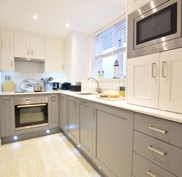 A kitchen with gray lower cabinets white upper cabinets an oven microwave a sink and a window at The Smokehouse Cottage in Southwold