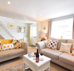 A living room with two sofas, colorful cushions, a white coffee table with wine and glasses, and a staircase in the background at Satis Cottage, Westleton