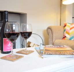A table with two glasses of red wine a bottle of wine magazines a beige sofa with colorful cushions and a fireplace in the background at Satis Cottage Westleton