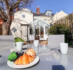An outdoor patio with a glass table holding a plate of croissants and grapes and three white mugs at Albion House Southwold