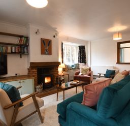 A living room with a teal sofa pink and teal cushions armchairs a wooden coffee table a fireplace with a fire a window seat with cushions and shelves with books at Rosedene Rickinghall