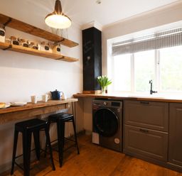A kitchen with wooden shelves jars cups two black stools and a washing machine under a window at Rosedene Rickinghall