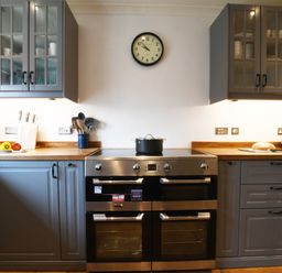 A kitchen with a stove and oven in the center a clock on the wall and gray cabinets with a toaster and knives on the wooden countertops at Rosedene Rickinghall