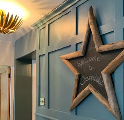 A blue paneled wall with a star-shaped chalkboard sign that says welcome to Cragside with a wooden door and a ceiling light fixture at Cragside Aldeburgh
