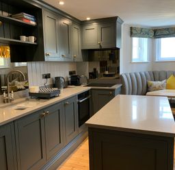 A kitchen with gray cabinets a countertop island a built in oven a curved bench seat and a dining table at Cragside  Aldeburgh