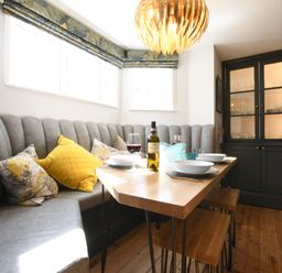 A dining area with a wooden table set with plates and wine glasses next to a curved gray banquette and a dark cabinet at Cragside Aldeburgh