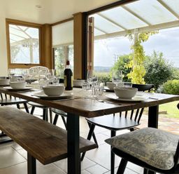 A dining area with a wooden table set with bowls glasses and a bottle and black chairs and a bench near open glass doors at Skelder Parham in Parham Near Framlingham