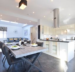 A dining table set with plates and wine glasses next to a kitchen with white cabinets and a stainless steel range hood at Pier View Southwold