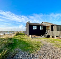 A black wooden building near a dock with boats under a blue sky at The Old Fisherman's Hut in Southwold