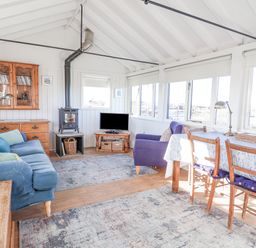 A living room with blue sofa wooden dining table and chairs wood stove and windows at The Old Fisherman s Hut in Southwold