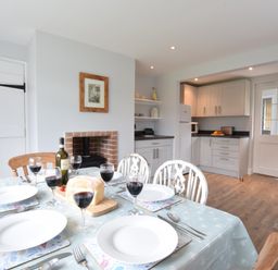 A dining table set with plates and wine glasses in a kitchen with white cabinets and a brick fireplace at Hoist Cottage Walberswick