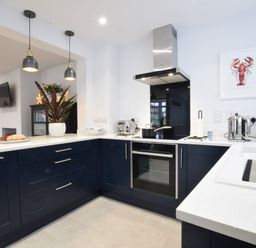A kitchen with blue cabinets, white countertops, hanging pendant lights, a stove with a pot, a toaster, a kettle, a coffee machine, and a framed lobster picture at Sea Green Cottage Walberswick