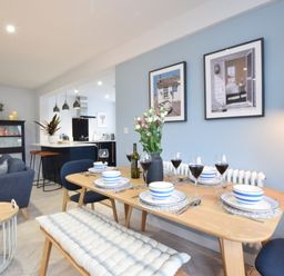 A dining area with a wooden table set for six and a kitchen with bar stools in the background at Sea Green Cottage Walberswick