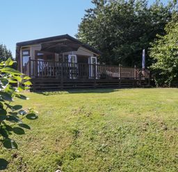 An outdoor view of a cabin with a wooden deck and patio umbrella surrounded by grass and trees at The Hide Out in Aldeburgh