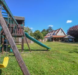 A garden with a children's playset that has a yellow swing and a green slide next to a house with a red tiled roof and a covered terrace at Moat Barn Hasketon Near Woodbridge