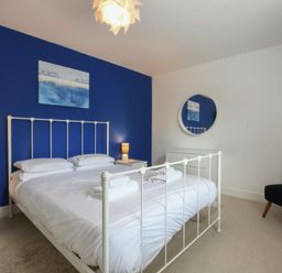 A bedroom with a white metal bed frame a blue accent wall two bedside tables with lamps a round wall mirror and a dark chair at The Gallery Blythburgh near Southwold