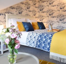 A bedroom with a bed with blue and yellow pillows and a yellow blanket a bedside table with a lamp and a vase with flowers on a round table at Driftwood Southwold