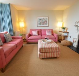 A living room with pink sofas a plaid ottoman a fireplace and blue curtains at Tamarisk in Aldeburgh