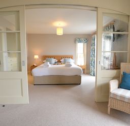 A bedroom with a large bed and side tables seen through sliding glass doors with a wicker chair in the foreground at Tamarisk Aldeburgh in Aldeburgh
