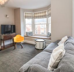 A living room with a gray sofa and white pillows a yellow chair a drum used as a table a bay window with striped blinds and patterned cushions a television on a wooden table at Osprey in Southwold