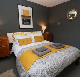 A bedroom with a double bed yellow cushions bedside tables lamps and a framed picture on the wall at Kestrel in Southwold
