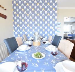 A dining table with plates wine glasses and a plate of asparagus in a room with patterned wallpaper and kitchen visible at St Marys View Southwold