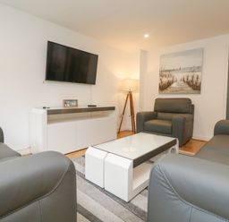 A living room with gray armchairs a white coffee table a wooden floor lamp a flat screen TV on the wall and a painting at Bluebell Retreat in Lostwithiel