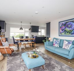 A living room and kitchen area with a blue sofa and ottoman a brown leather armchair a dining table with chairs and bench a telescope and floral painting at Watlands Mynytho near Abersoch