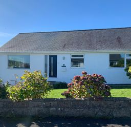 A single-story white house with a dark door and three windows a garden with bushes and flowers in front at Watlands Mynytho near Abersoch