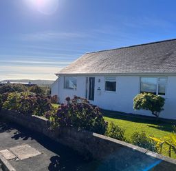 A white house with a slate roof and a garden with bushes and a small tree overlooking a coastal landscape at Watlands in Mynytho near Abersoch