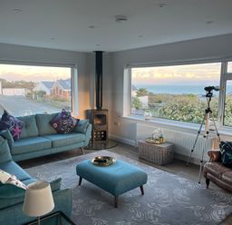 A living room with blue sofas and an armchair with large windows showing a sea view at Watlands in Mynytho near Abersoch