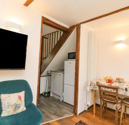 A room with a green chair with a rooster cushion a wall-mounted tv a small dining table with two chairs and a kitchenette under stairs at Tiddlers Cottage in Uploders near Bridport