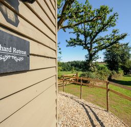The side of a building with a sign reading Orchard Retreat and a fenced grassy area with trees at Orchard Retreat East Worlington near Witheridge