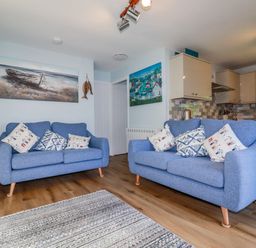 A living room with two blue sofas and cushions with a kitchen area in the background at Woodside in Rosecraddoc near St Cleer