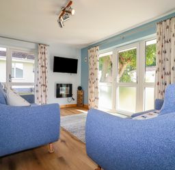 A living room with two blue armchairs a wall-mounted television and large windows with curtains at Woodside Rosecraddoc near St Cleer