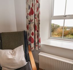 A corner with a chair with cushions next to a window with patterned curtains and a radiator below at Ceunant near Newport