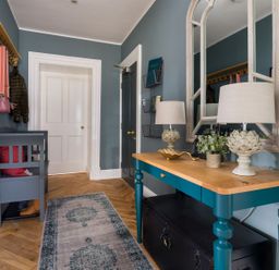An entryway with a blue bench, a wooden table with lamps and plants, a large mirror, a coat rack, and a rug on a wooden floor at Wansfell at Lipwood House in Windermere