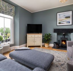 A living room with a blue sofa and ottoman a television on a cabinet a fireplace and a window with a potted plant at Wansfell at Lipwood House Windermere