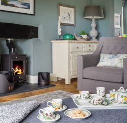 A living room with a burning stove a gray armchair and a tea set on a table at Wansfell at Lipwood House in Windermere
