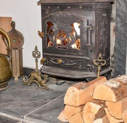 A close-up of a wood-burning stove with fire inside with firewood stacked nearby and brass fireplace tools at Naunton Cottage in Lyme Regis