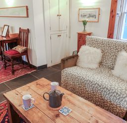 A living room with a wooden coffee table with a teapot and cups a patterned sofa with cushions wooden chairs and a table against the wall at Naunton Cottage in Lyme Regis