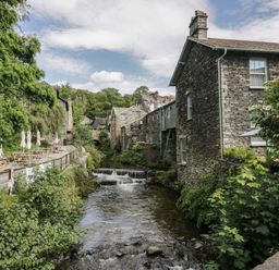 A river flowing between stone buildings and trees with picnic tables along the river at Rose Cottage Ambleside