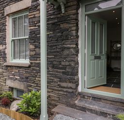 A stone exterior wall with a green framed window and an open green door showing a glimpse of an interior with tiled floor at Rose Cottage in Ambleside
