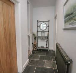 A hallway with a wooden door to the left a radiator on the right and a small grey hall stand with a round mirror and umbrella at Rose Cottage Ambleside