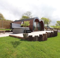 A yard with green grass a wooden patio with a hot tub a potted tree and a wooden pod building with round windows at Creuddyn in Llanfihangel-y-Creuddyn near Aberystwyth