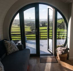 A seating area with a sofa and pillows next to a round glass door opening to a balcony with a view of green fields at Creuddyn in Llanfihangel-y-Creuddyn near Aberystwyth